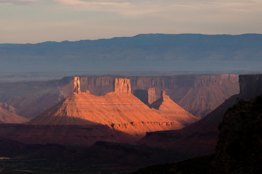 Sunlight illuminating towering sandstone formations in a vast canyon landscape with distant mountain range under a soft sky